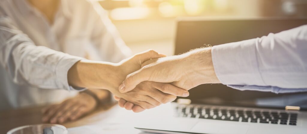 Two people shaking hands across a desk with a laptop and papers, suggesting a successful business meeting or agreement. The background is softly lit, creating a professional atmosphere.