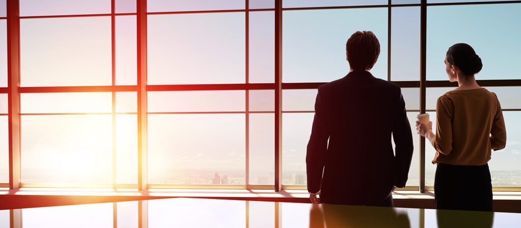 Two people in business attire stand by large office windows, looking out at a cityscape during sunrise or sunset; one holds a coffee cup.