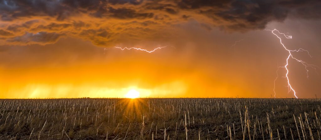 A dramatic sunset lights up the sky with orange and yellow hues over a harvested field, while dark clouds and multiple lightning bolts strike in the distance.