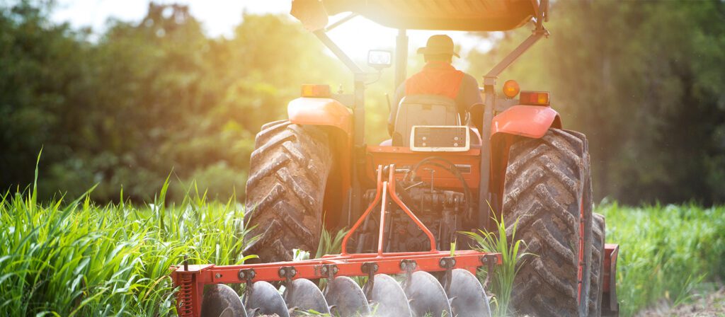 A farmer drives a red tractor through a green field on a sunny day, cultivating the soil with attached agricultural equipment. Tall grass and trees are visible in the background.