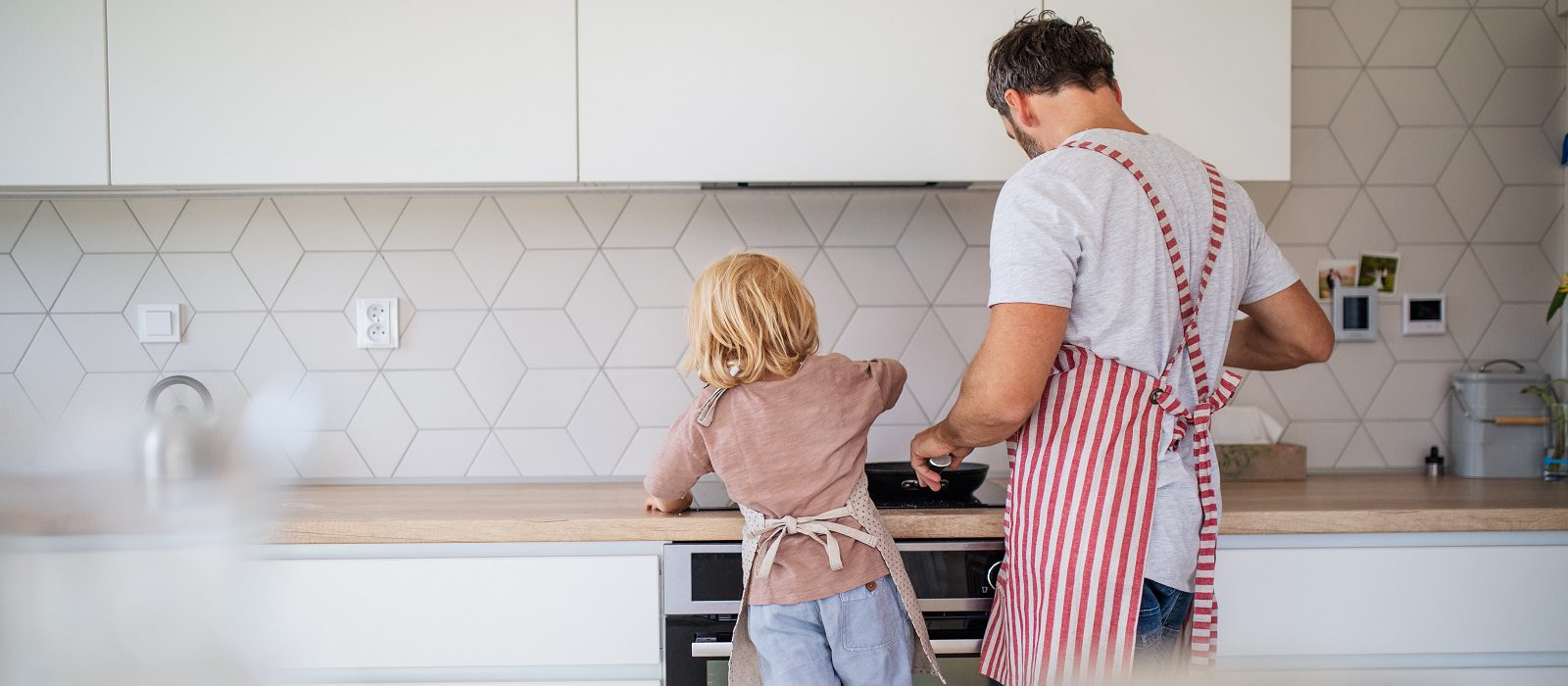 A rear view of small boy helping father indoors in kitchen with making pancakes.