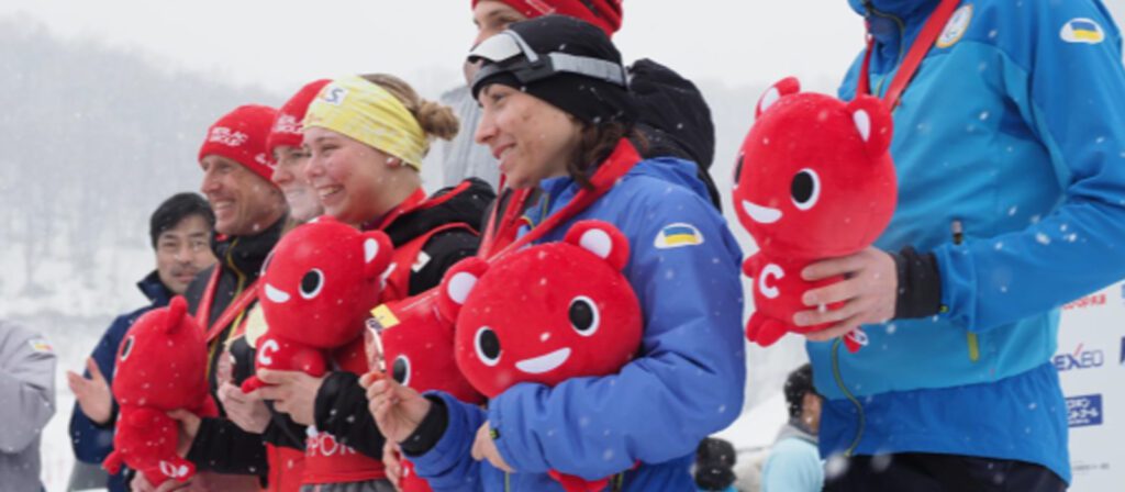 A group of athletes stand outdoors in the snow, smiling and holding red mascot plush toys and medals. They are dressed in winter sports gear and appear to be celebrating on a podium.