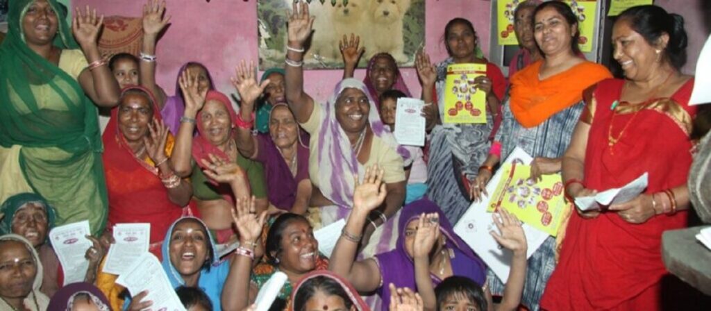 A group of women, dressed in colorful traditional clothing, sit and stand closely together indoors, many smiling and waving while holding papers and booklets, appearing joyful and engaged in a community gathering.