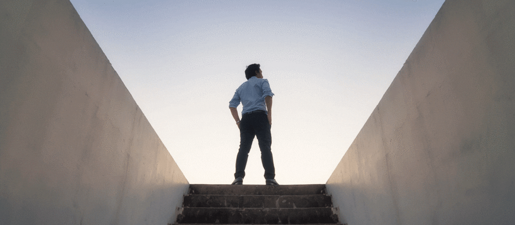 A man stands at the top of a staircase between two concrete walls, looking up at the sky, with his hands on his hips. The scene is lit by soft natural light, creating a sense of ambition and possibility.