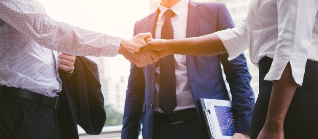 Three businesspeople in professional attire stand outside, two of them shaking hands while the third observes. One holds a tablet displaying a graph. The scene suggests a successful business agreement or partnership.