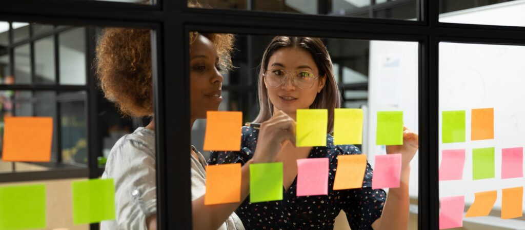 Two women stand behind a glass wall covered with colorful sticky notes, discussing ideas and pointing at the notes in a modern office setting.