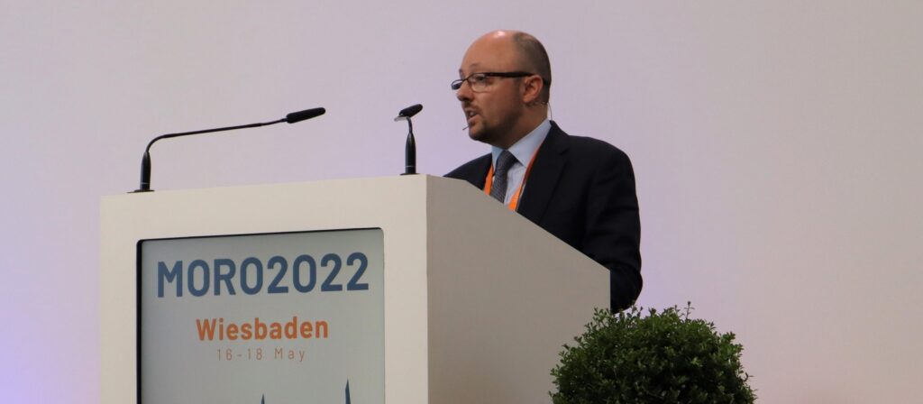 A man in a suit and tie speaks at a white podium with microphones. The podium displays MORO2022 Wiesbaden 16-18 May on the front. There is a plant in the foreground and a plain light background.