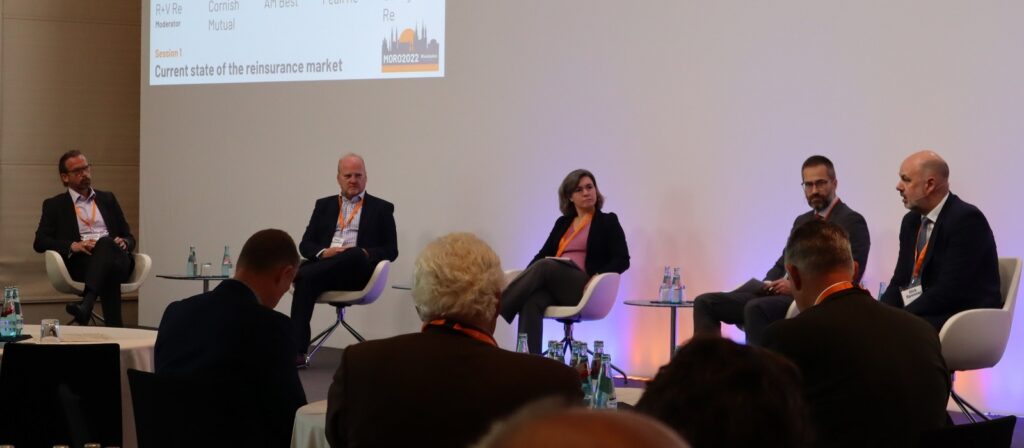 Five panelists sit on stage in chairs, engaged in discussion at a conference. An audience sits in front of them. A projected slide behind them reads Current state of the reinsurance market.