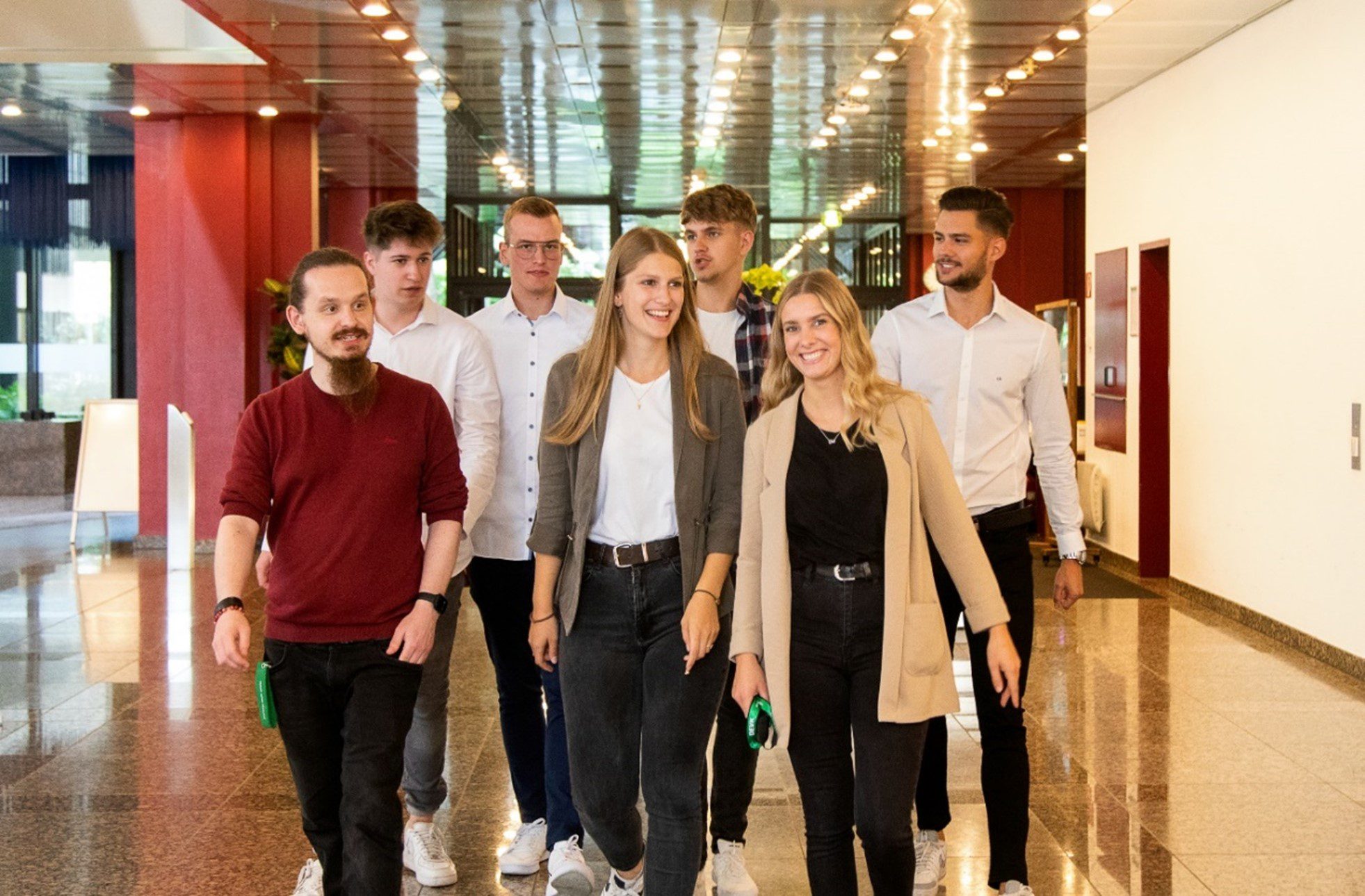 Eight young adults, casually dressed, walk together in a brightly lit hallway with red walls and reflective floors, smiling and appearing cheerful.