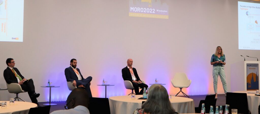 Four people sit on a stage with microphones and water bottles during a conference panel, while one woman stands and presents near a large projection screen. The event is labeled “MOR2022 Wiesbaden.”.