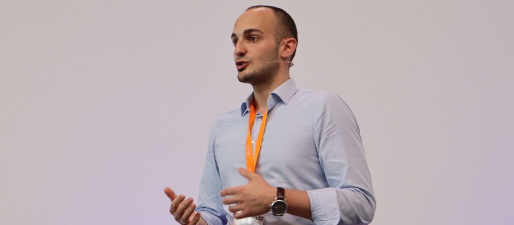 A man wearing a light blue shirt and orange lanyard speaks on stage with a microphone headset, gesturing with his hands against a plain light background.