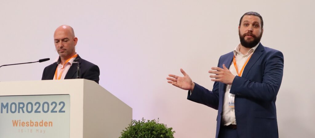 Two men in suits speak at a podium labeled MORO2022 Wiesbaden 16-18 May; one stands behind the podium looking down, while the other gestures with his hand, facing the audience.
