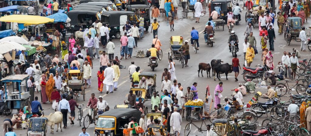 A busy street scene in India with people walking, vendors selling goods, auto rickshaws, bicycles, street stalls, and cows moving through the crowd in a lively, colorful environment.
