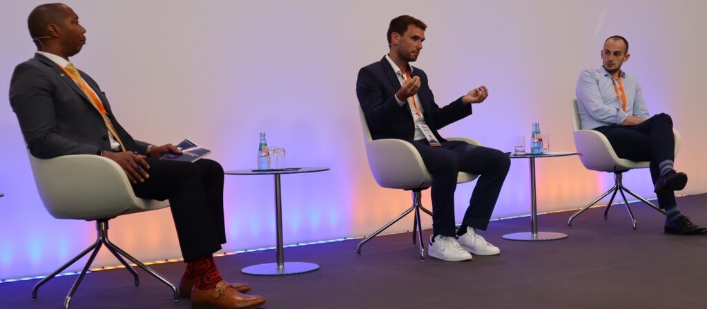 Three men sit spaced apart on modern white chairs, engaged in a panel discussion. Each has a bottle of water on a small table beside them. The background is plain with soft orange lighting.