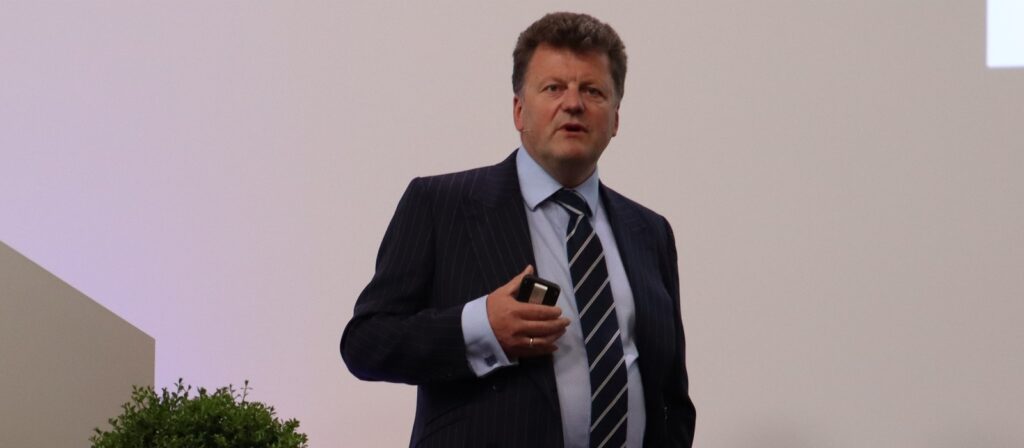 A man in a dark pinstripe suit and striped tie is standing and speaking, holding a small device. There is a green plant to his left and a plain light background behind him.