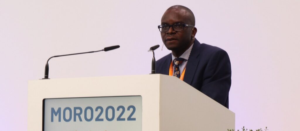 A man in a suit and glasses stands at a podium with two microphones, speaking at an event labeled MORO2022 against a plain white background.