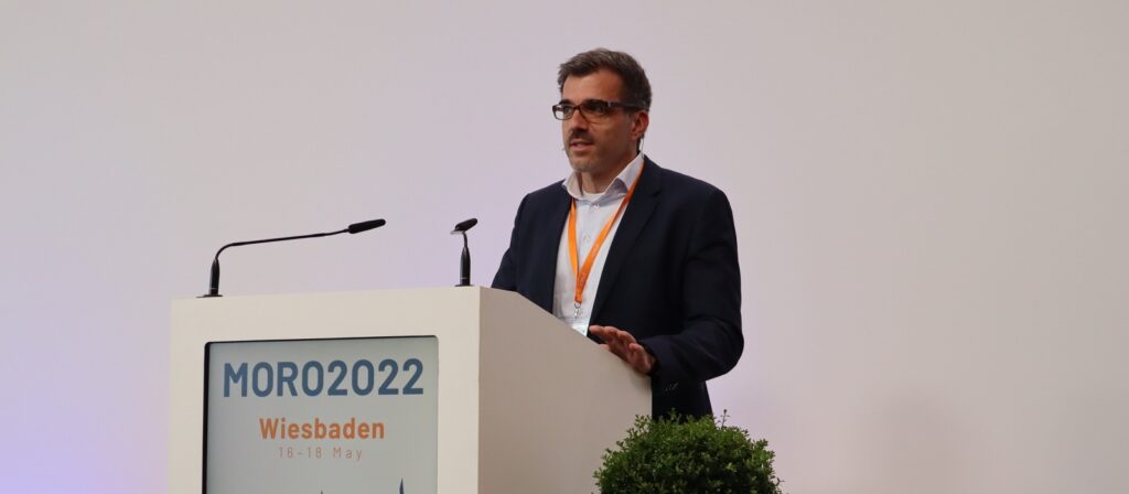 A man in a suit and glasses speaks at a podium labeled MORO2022 Wiesbaden 16-18 May with two microphones, against a plain background and a small green plant in front.