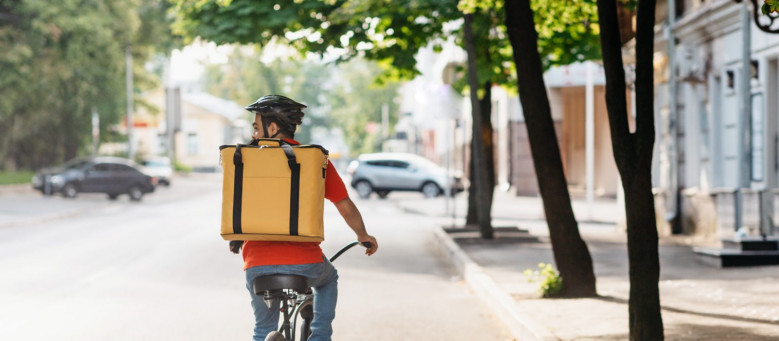 Courier on bicycle with parcel, bike, delivery service. Young guy in uniform, protective helmet with yellow thermo bag riders on road in city in summer and searches client address, empty space, back