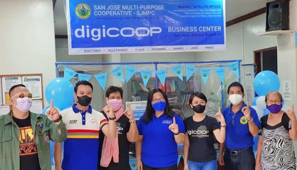 Seven people wearing face masks stand in a row, some in blue shirts, in front of a Digicoop Business Center banner with blue balloons and bunting. They are holding up one finger, posing for a group photo.