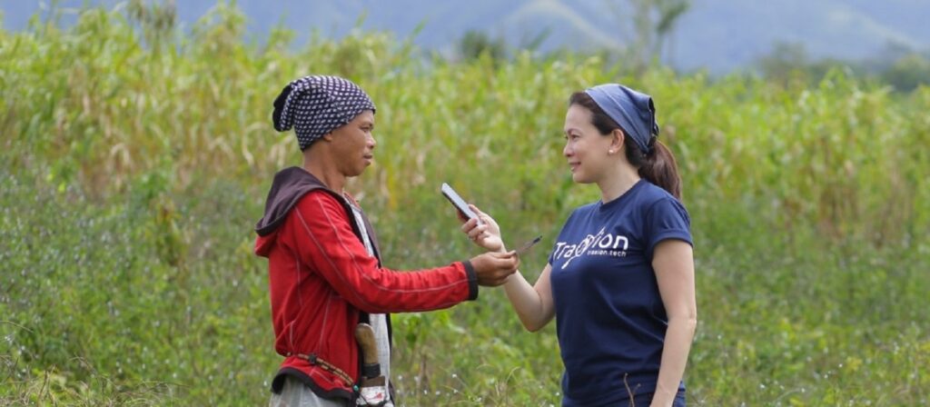 A woman in a navy blue shirt interviews a man in a red jacket using a smartphone, standing in a green field with mountains in the background.