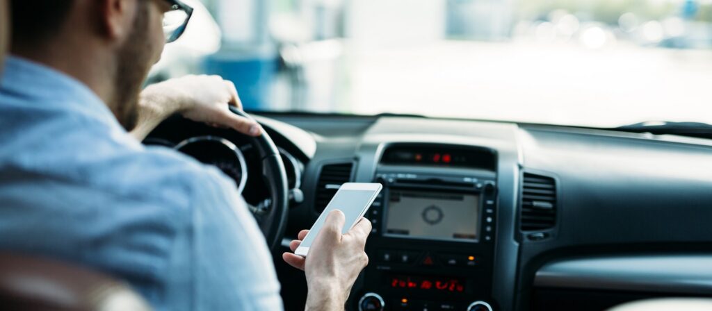 A man sits in the drivers seat of a car, holding the steering wheel with one hand while looking at a smartphone in his other hand. The dashboard and in-car navigation system are visible.