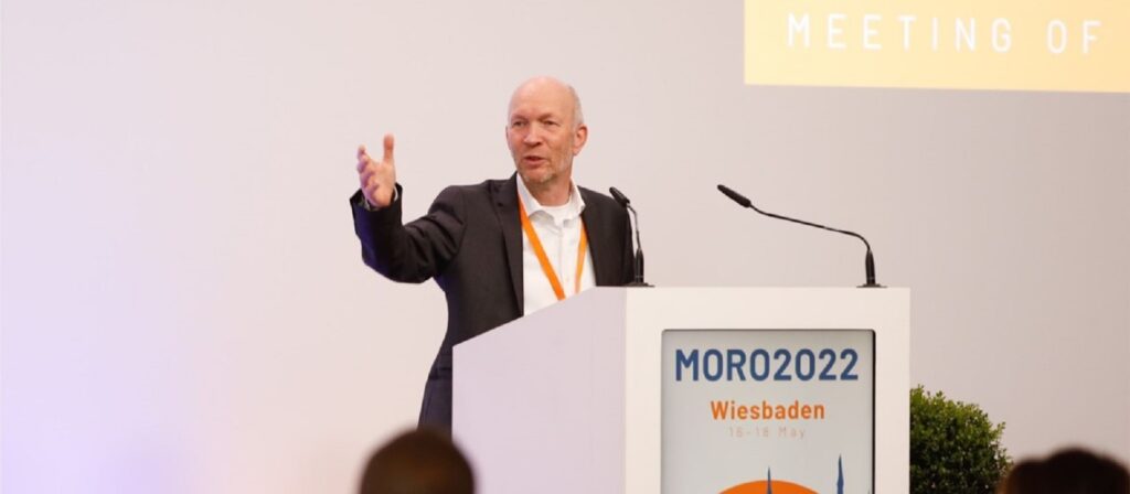 A man in a suit speaks at a podium labeled MORO2022 Wiesbaden 16-18 May during a conference. He gestures with one hand, with a microphone and a projection screen behind him.
