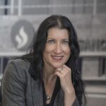 A woman with long black hair and blue eyes, wearing a gray blazer and a necklace, smiles while resting her chin on her hand. The background is blurred and appears to be an indoor office setting.