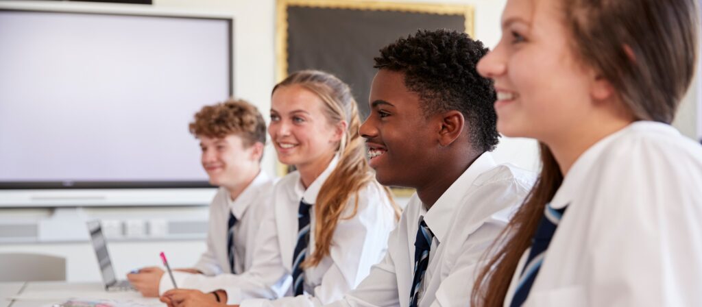 Four high school students in uniform sit at desks in a classroom, smiling and looking toward the front of the room. A whiteboard and large monitor are visible in the background.