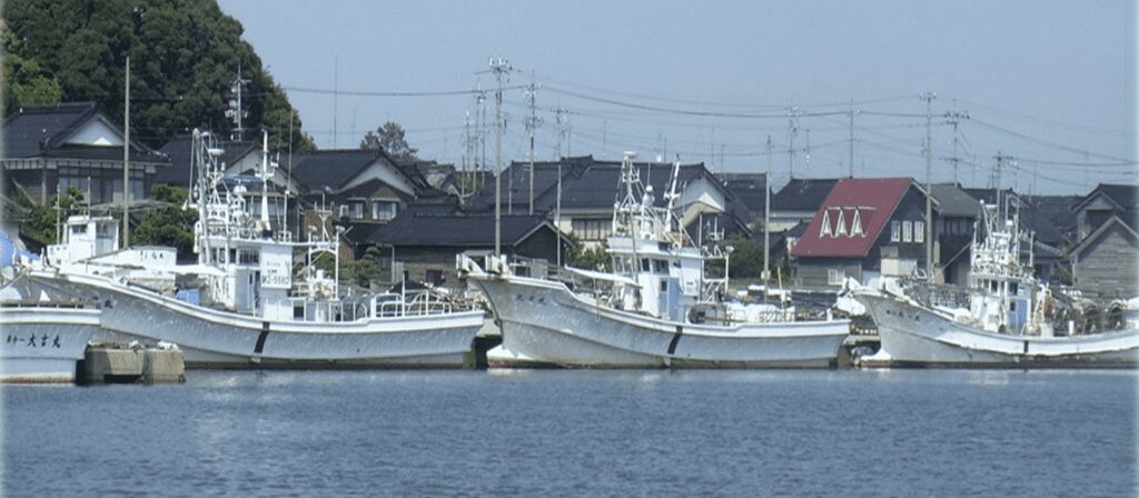 Several white fishing boats are docked along a calm waterfront, with traditional Japanese houses, trees, and utility poles visible in the background under a clear sky.