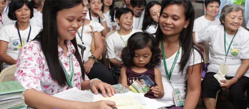 A smiling woman signs documents at a table, while another woman holding a young girl sits beside her. Several women in white shirts are seated in the background, watching the interaction.