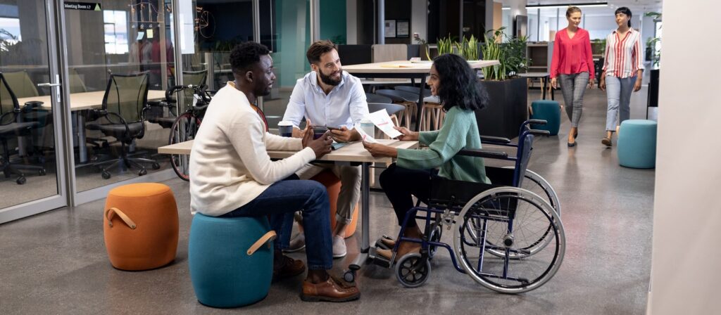 Three colleagues, including a woman in a wheelchair, sit around a table having a discussion in a modern office. Two women walk in the background. The setting is open and inclusive.
