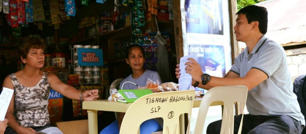 Three people sit around a yellow table outside a small store, engaged in conversation. Snacks and papers are on the table. Shelves with various goods are visible in the background.
