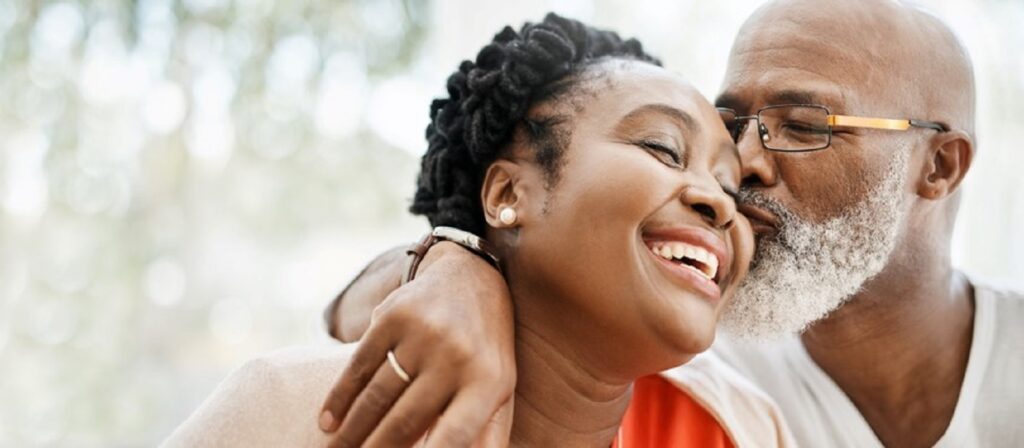 An older man with glasses lovingly kisses a smiling woman on the cheek, both appearing joyful and relaxed in a close embrace, with a soft, blurred background.