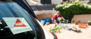 A close-up of a car window with a MAIF sticker, parked near a garden where a person is tending to plants in the background. The scene is bright and outdoors.