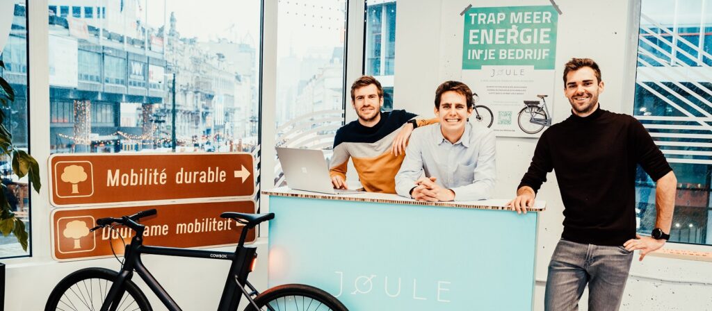 Three smiling men stand behind a counter with a “JOULE” logo next to a black bicycle. Signs promoting sustainable mobility and energy are on the wall and windows overlook a busy city street.