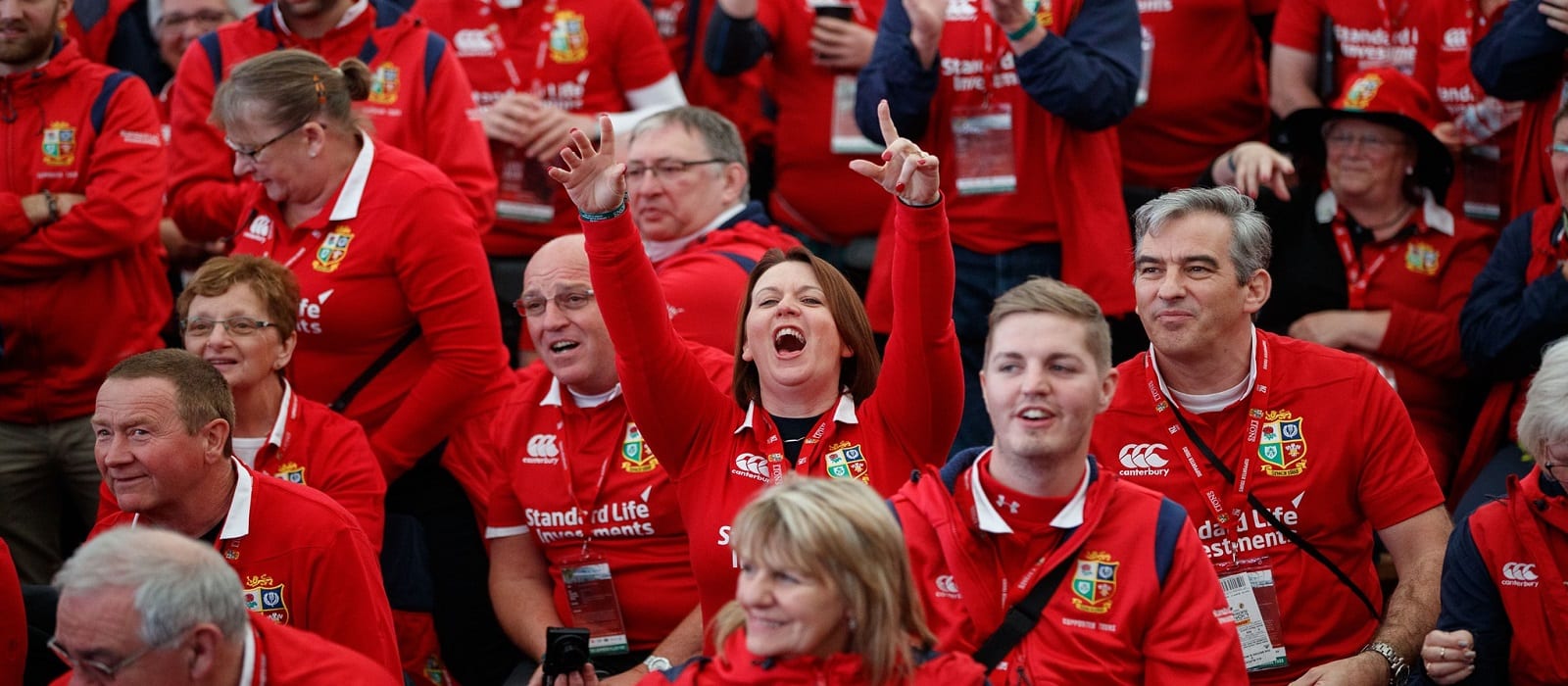 A crowd of fans wearing matching red rugby shirts sits closely together. A woman in the center raises both arms and cheers enthusiastically, surrounded by smiling and attentive people.