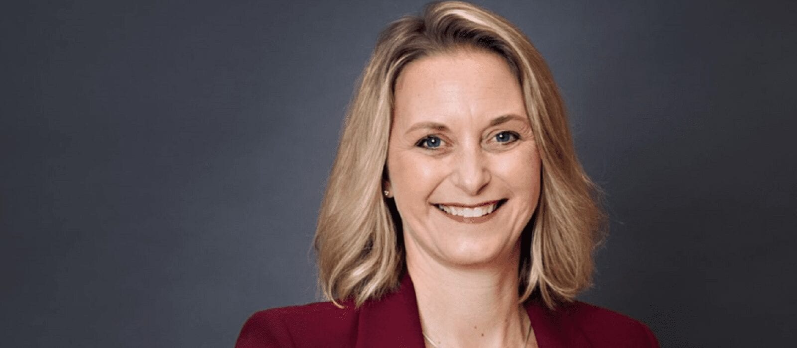 A woman with shoulder-length blonde hair smiles at the camera. She is wearing a maroon blazer over a light top and is posed in front of a plain dark gray background.