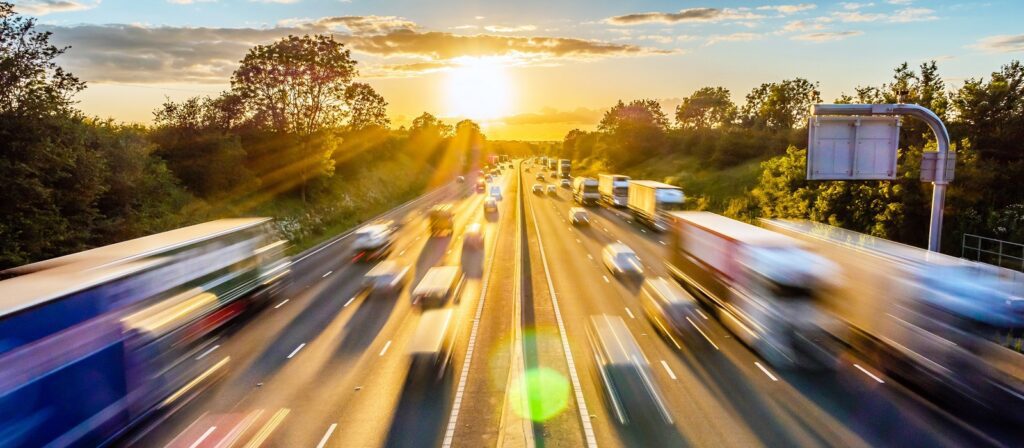 A busy highway at sunset shows blurred cars and trucks speeding in both directions, with sunlight streaming through trees on either side of the road.