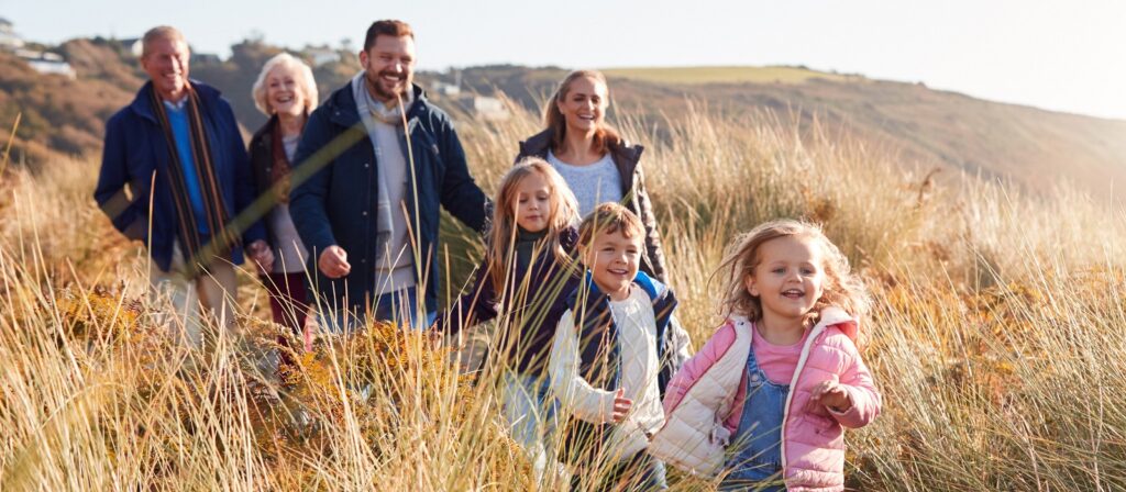 Three generations of a family, including children and adults, walk happily through tall grass on a sunny day, with hills visible in the background.