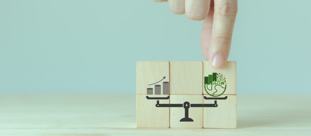A hand presses on wooden blocks showing a scale balancing money and an upward arrow on one side with a green globe and leaves on the other, symbolizing financial and environmental balance.
