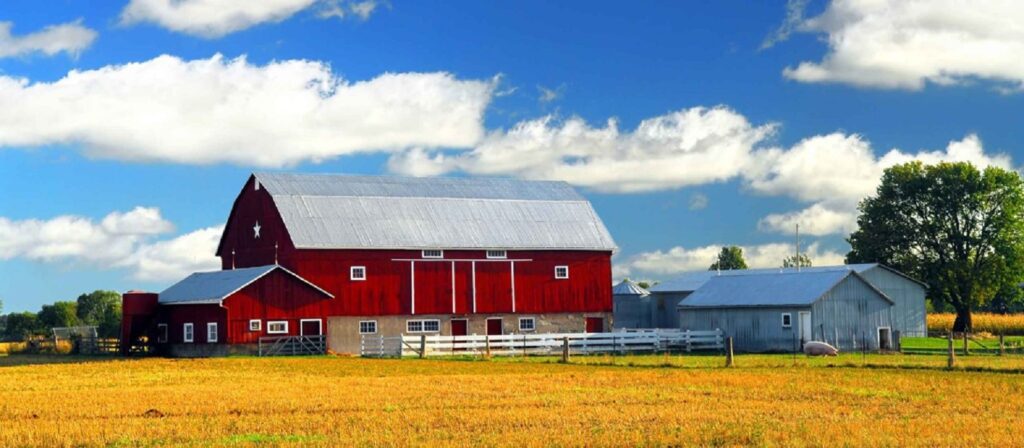 A large red barn with a white roof stands beside a smaller gray building, surrounded by a white fence and open fields under a bright blue sky with scattered clouds. Trees and farmland are visible in the background.