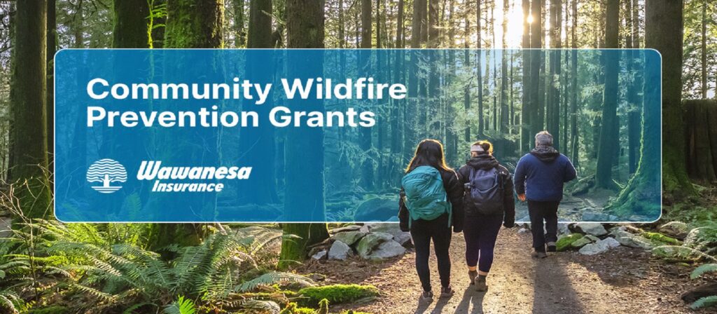 Four people walk on a forest trail surrounded by tall trees and sunlight. A blue banner reads: Community Wildfire Prevention Grants, Wawanesa Insurance.