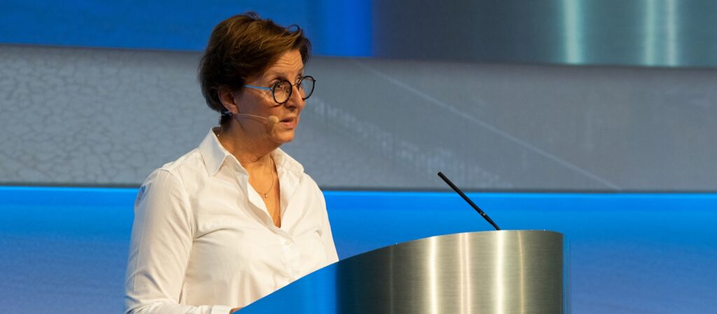 A woman wearing glasses and a white shirt speaks at a podium with a microphone, against a blue and metallic background.