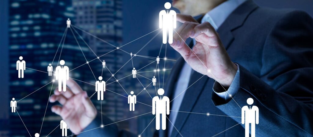 A person in a suit interacts with a virtual network of connected human icons, representing digital communication or social networking, with a cityscape blurred in the background.