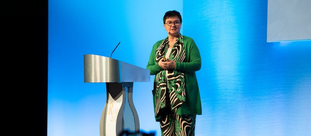 A woman in a green patterned outfit stands on stage at a podium, speaking to an audience with a blue background behind her.
