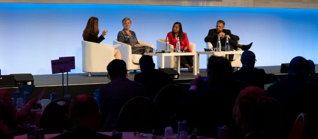 Four people are seated on a stage in a panel discussion, facing an audience. Three women and one man sit in white chairs with small tables between them, speaking and listening under blue lighting.