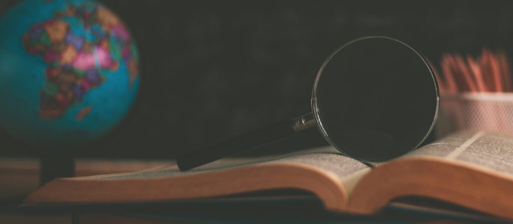 A magnifying glass rests on an open book, with a globe and a pencil holder visible in the blurred background, suggesting a study or research setting.