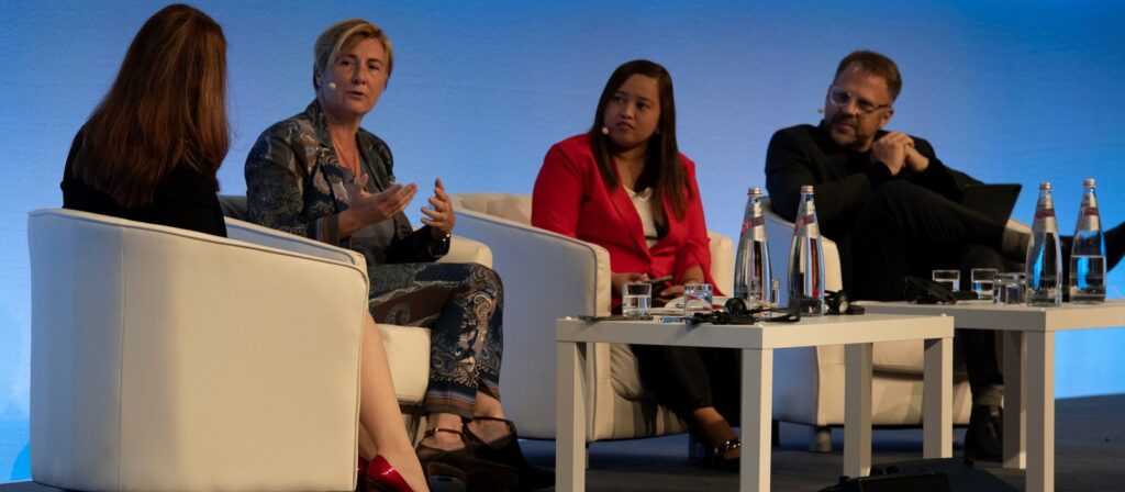 Four people sit on white chairs on a stage, engaged in a panel discussion. Two women and one man face the audience, while another woman sits with her back to the camera. Bottled water and glasses are on tables in front of them.