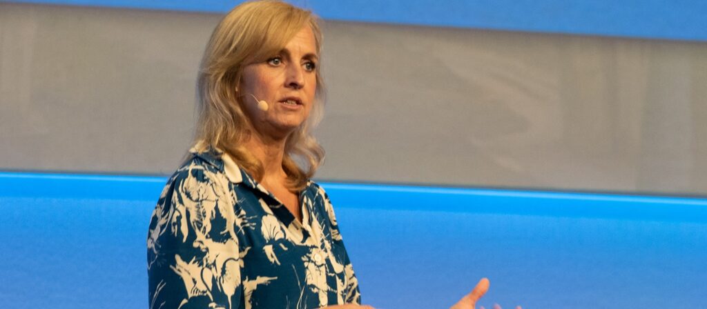 A woman with long blonde hair, wearing a blue and white patterned blouse and a headset microphone, speaks on stage against a blue and gray background.