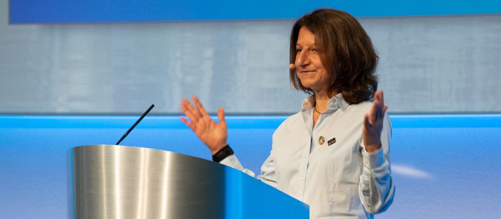 A woman with long brown hair speaks at a podium with a microphone headset, gesturing with both hands raised. She is wearing a light blue shirt and standing in front of a blue and white background.